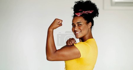 Happy Confident young African American woman showing strength, flexing arm looking at camera. One black latina shows muscle, we can do it symbol, wearing yellow shirt puzzle 716020282