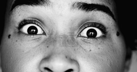 One scared young black woman macro close-up eyes looking directly at camera in SHOCK and HORROR feeling worry and paranoia to scary revelation, eye wide open