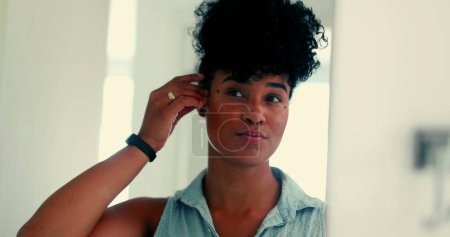 Téléchargez les photos : Une jeune femme brésilienne noire heureuse ajustant sa coiffure devant un miroir se préparant à sortir. Élégant afro-américain 20s adulte fille souriant se sentant confiant - en image libre de droit