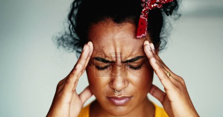 Foto de Mujer frustrada joven cara de cerca que tiene un dolor de cabeza poniendo las manos en las sienes laterales sintiendo presión mental. Estresado afroamericano 20s persona - Imagen libre de derechos
