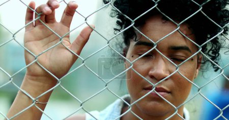 Une jeune femme noire sérieuse derrière une clôture métallique, symbolisant l'isolement en crise, penchant la main et regardant la caméra avec une expression solennelle