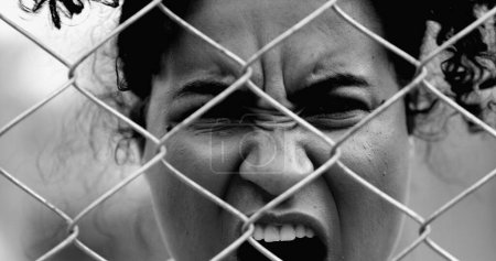 Desperate Young African American woman feeling outrage screaming with rage behind metal fence barrier frowning and open mouth screaming in anger