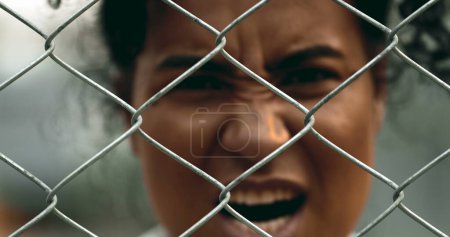 Photo for One upset young black woman yelling behind metal fence barrier looking at camera grimacing in anger and screaming in despair. 20s person feeling outrage - Royalty Free Image