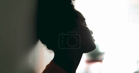 Foto de Triste joven afroamericana reflexiva se apoya en la pared mirando a la ventana con expresión pensativa melancólica luchando contra la depresión durante tiempos difíciles - Imagen libre de derechos