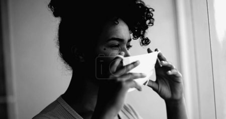 Foto de Una joven mujer negra poniendo máscara facial covid atascado en casa en intenso retrato monocromático retrato cara de cerca mirando a la ventana desde el apartamento - Imagen libre de derechos