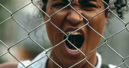Photo for One upset young black woman yelling behind metal fence barrier looking at camera grimacing in anger and screaming in despair. 20s person feeling outrage - Royalty Free Image
