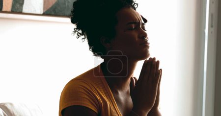 Foto de Una joven religiosa negra orando tranquilamente en casa buscando ayuda y apoyo de Dios con los ojos cerrados y señalando al cielo sintiendo fe y espiritualidad - Imagen libre de derechos