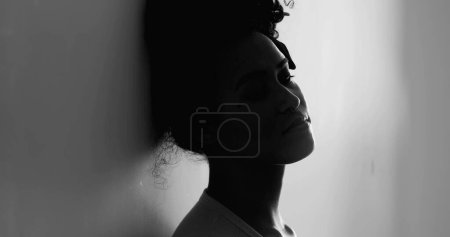 Foto de Una joven mujer negra deprimida apoyada en la pared sintiéndose sola en una tranquila desesperación luchando contra una enfermedad mental. Silueta de una persona afroamericana de 20 años que sufre sola en blanco y negro - Imagen libre de derechos