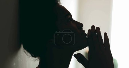 One spiritual young black woman PRAYING to GOD leaning on wall in profile silhouette. Person seeking help and support during difficult times with hands clenched looking up with HOPE