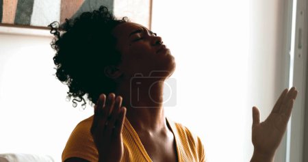 Eine religiöse junge schwarze Frau betet leise zu Hause und sucht Hilfe und Unterstützung bei Gott mit geschlossenen Augen und zeigt auf den Himmel, der Glauben und spirituelle Gefühle empfindet