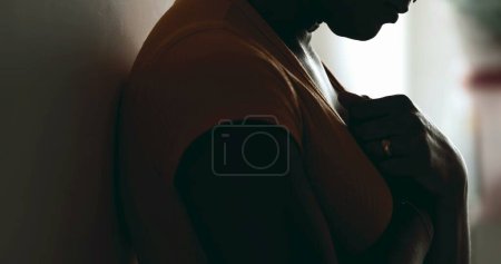 Photo for Young African American woman struggling during challenging times looking down with hands on chest seeking help and support in silhouette. Profile of a young black latina seeking help - Royalty Free Image