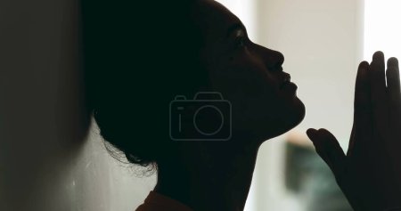 Photo for One spiritual young black woman PRAYING to GOD leaning on wall in profile silhouette. Person seeking help and support during difficult times with hands clenched looking up with HOPE - Royalty Free Image