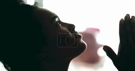 Photo for One spiritual young black woman looking up at sky in PRAYER feeling hopeful and faithful to a higher power. African American 20s person seeking God's support and help, profile close-up - Royalty Free Image