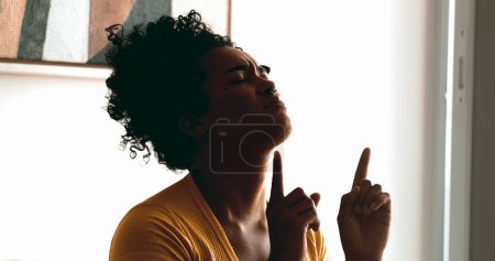 Eine religiöse junge schwarze Frau betet leise zu Hause und sucht Hilfe und Unterstützung bei Gott mit geschlossenen Augen und zeigt auf den Himmel, der Glauben und spirituelle Gefühle empfindet