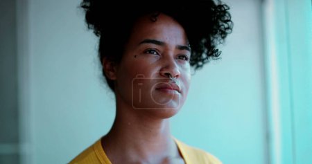 Photo for One pensive young black woman gazing from apartment balcony window close-up face. African American 20s person with curly and stern serious expression with contemplative emotion - Royalty Free Image