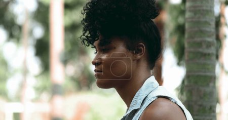 Téléchargez les photos : Une jeune femme noire contemplative présente un visage en gros plan dans une profonde réflexion mentale assise à l'extérieur du parc avec une émotion méditative solennelle - en image libre de droit