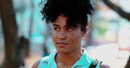 Foto de Retrato de una joven mujer negra pensativa de pie al aire libre en estado meditativo pacífico, cara de cerca en el seguimiento de disparo durante el día soleado y la emoción contemplativa - Imagen libre de derechos