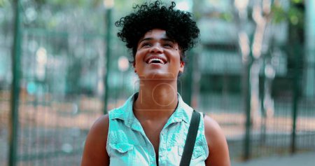 Foto de Una joven negra despreocupada mirando al cielo sonriendo sintiendo alegría durante el soleado día del parque parada afuera - Imagen libre de derechos