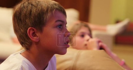 Téléchargez les photos : Candid jeune garçon regarder écran de télévision film vie réelle heureux authentiques réactions des enfants - en image libre de droit