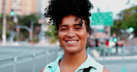 Foto de Una joven negra feliz de ascendencia africana con el pelo rizado de pie en el entorno urbano sonriendo. Retrato de una chica de 20 años adulta sudamericana - Imagen libre de derechos