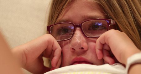 Photo for Closeup of little girl face watching scary movie screen child wearing glasses paying attention to story - Royalty Free Image