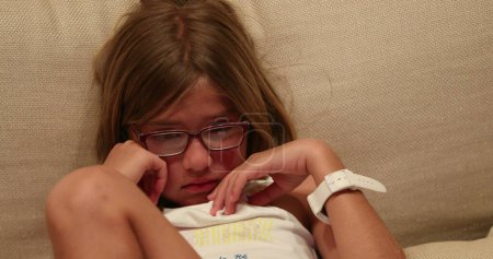 Téléchargez les photos : Petite fille portant des lunettes de lecture assise sur le canapé regardant un film effrayant - en image libre de droit