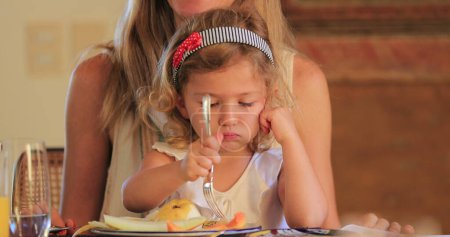 Little girl eating lunch holding fork on mother lap candid child and mom