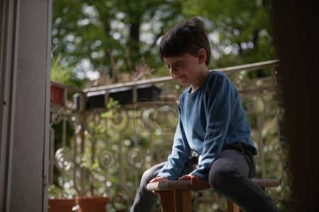 Téléchargez les photos : Un jeune garçon en chemise bleue est assis sur un petit banc en bois à l'extérieur sur un porche. Sa tête est légèrement inclinée, et il semble perdu dans la pensée, avec une toile de fond de verdure ajoutant une touche sereine. - en image libre de droit