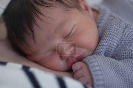 Photo for A close-up of a newborn baby peacefully sleeping with its tiny hand nestled near its face. The baby wears a cozy gray knitted sweater, highlighting its delicate and serene features, including fine, dark hair. - Royalty Free Image