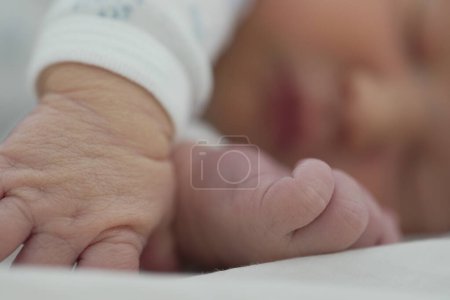 El recién nacido está durmiendo tranquilamente, con un primer plano de la cara y la mano descansando cerca. El enfoque suave en el fondo enfatiza el sueño calmado y sereno del bebé, vestido con un onesie blanco con patrones delicados.