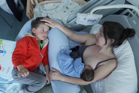 Eine Mutter liegt in einem Krankenhausbett und berührt sanft den Kopf ihres älteren Kindes, das neben ihr liegt, während sie ihr in eine blaue Decke gehülltes Neugeborenes stillt. Die Szene strahlt Wärme und familiäre Verbundenheit aus.