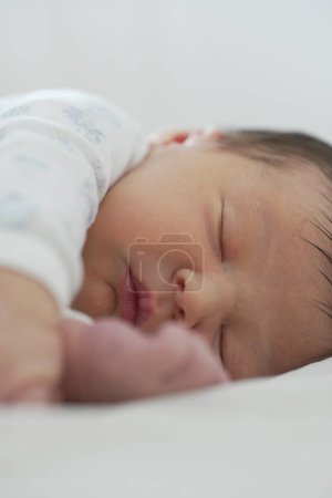 El recién nacido está dormido sobre una manta blanca, con un brazo extendido hacia adelante y la cara ligeramente hacia un lado. La luz suave y la expresión serena capturan la esencia pura y pacífica del descanso del bebé.