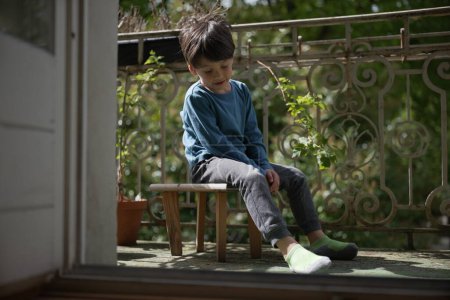 Téléchargez les photos : Un jeune garçon en chemise bleue est assis sur un petit banc en bois à l'extérieur sur un porche. Il semble pensif, regardant vers le bas avec une expression réfléchie, entouré de plantes vertes et de lumière naturelle. - en image libre de droit