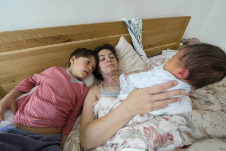 Mother resting in bed with her newborn baby and older child, sharing a tender moment, showcasing the bond and love between siblings and their mother in a cozy, family setting