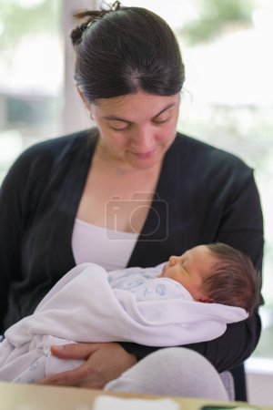 Foto de Madre joven sosteniendo a su bebé recién nacido envuelto en una manta suave, mirando hacia abajo con amor y ternura, enfatizando el fuerte vínculo maternal y el ambiente pacífico y nutritivo - Imagen libre de derechos
