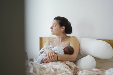Téléchargez les photos : Mère allaitant son nouveau-né assis au lit, montrant un moment d'éducation, de soins maternels, et le lien intime formé pendant le processus d'allaitement dans un environnement calme à la maison - en image libre de droit