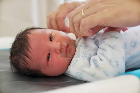 Neugeborenes Baby liegt auf einem Wickelkissen, bekleidet in einem weißen Einteiler mit blauen Abdrücken, die Hand ausgestreckt, mit zarten Fingern und feiner Hautstruktur, aufgenommen in einem sanft beleuchteten Innenraum