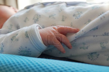 Foto de Un primer plano de la mano de un recién nacido agarrando suavemente el borde de un onesie modelado suave, blanco y azul, mostrando las características diminutas y delicadas de los dedos de un bebé y la textura de la tela acogedora - Imagen libre de derechos
