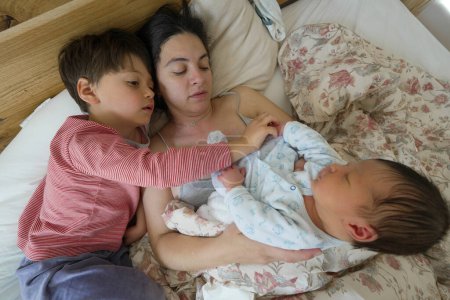 Mother cuddling her newborn baby in bed while her older child looks on, capturing a tender and loving family moment, emphasizing the close bond and affection among siblings and mother