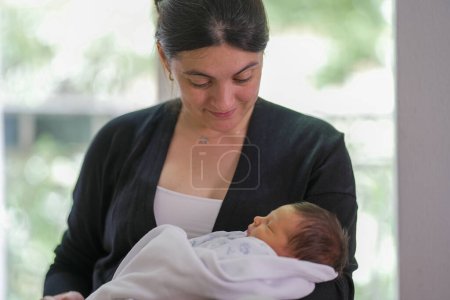 Foto de Madre sosteniendo a su bebé recién nacido envuelto en una manta suave, mirando hacia abajo con amor y ternura, enfatizando el fuerte vínculo maternal y el ambiente pacífico y nutritivo - Imagen libre de derechos
