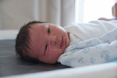 Neugeborenes Baby in einem weißen Onesie mit blauen Drucken, auf einer Wickelmatte liegend, mit wachem Gesichtsausdruck aufblickend, eingefangen in sanftem Licht, mit feinen Haardetails und Gesichtszügen