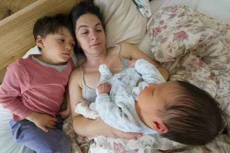 Mother holding her newborn baby in bed with her older child lying beside them, capturing a sweet family moment of bonding, highlighting the love and connection shared among them