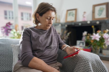Photo for Elderly woman with short hair in a grey shirt sitting on a patterned couch, looking at her red smartphone with a serious expression, surrounded by a cozy, well-decorated living room - Royalty Free Image