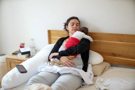 Foto de Madre descansando en una cama mientras sostiene a su bebé recién nacido que está vestido con pantalones rojos y un top blanco. La madre y el bebé están dormidos, mostrando un momento de descanso pacífico. - Imagen libre de derechos