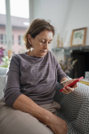 Photo for Senior woman sitting on a couch, holding a red smartphone and looking down thoughtfully, dressed in a grey shirt, with a warm, inviting living room featuring floral arrangements and soft lighting - Royalty Free Image