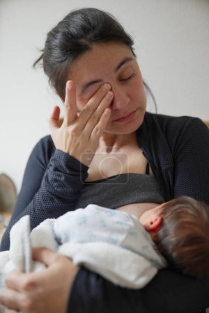 Mutter, erschöpft und emotional, wischt sich Tränen, während sie ihr Neugeborenes stillt. Dieses ergreifende Bild zeigt die Dualität von Freude und Müdigkeit in der frühen Mutterschaft und unterstreicht die Verbindung zwischen Mutter und Kind