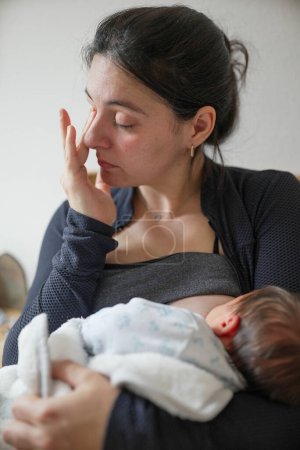 Nouvelle mère, essuyant ses yeux tout en allaitant son bébé, capturant l'épuisement émotionnel et physique de la maternité précoce. Une représentation sincère de l'amour et des défis auxquels font face les nouveaux parents