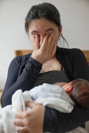 Neue Mutter wischt sich die Augen, während sie ihr Baby stillt, und zeigt den emotionalen und physischen Tribut der frühen Elternschaft. Erfasst die Intensität der Wochenbetterfahrungen und die Liebe, die sie mit ihrem Kind teilt