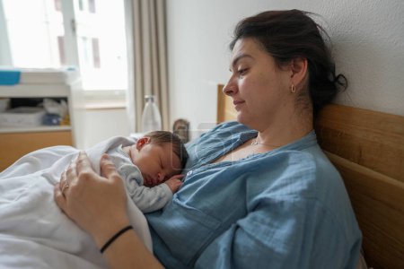 New mother resting in bed, holding her sleeping newborn baby close to her chest. The serene moment captures the deep bond between mother and child, highlighting the tranquility of early motherhood.