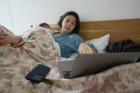 Photo for New mother multitasking in bed, using a laptop while her newborn sleeps on her chest. The image captures the balance between technology and parenting, showcasing the modern dynamics of motherhood. - Royalty Free Image